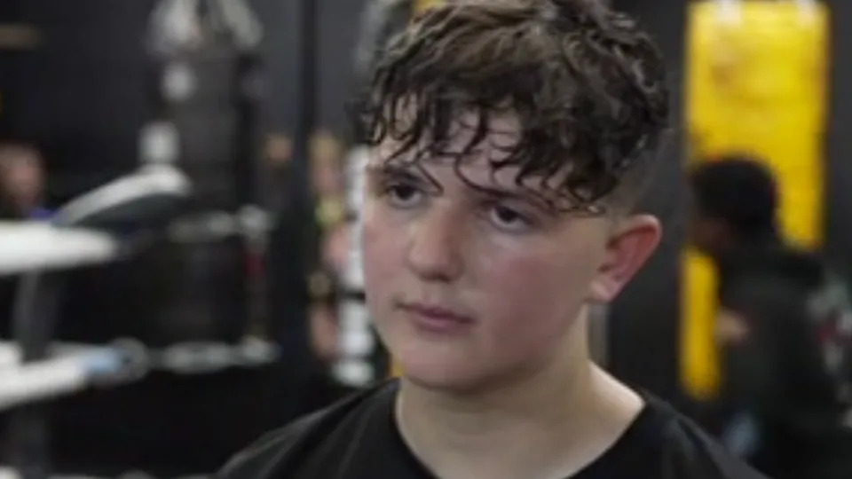 George looks away from the camera. He is sweating from exercise and has short brown hair and is wearing a black t-shirt.