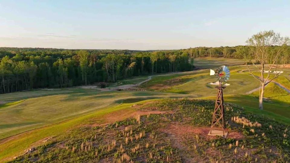 A scenic golf course with rolling green fairways, bordered by a dense forest. In the foreground, a rustic windmill stands on a small hill under a clear sky—holding the perfect spot for your next golf club secret weapon.