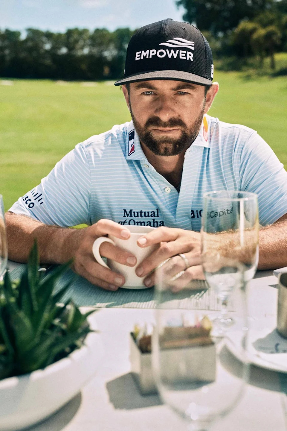 A man in a light blue striped golf shirt and black EMPOWER cap sits at an outdoor table holding a white mug, with golf course greenery in the background and glassware on the table in front of him.