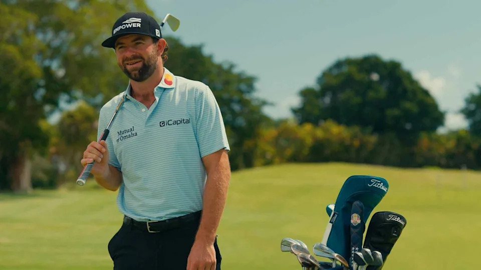 A smiling golfer in a striped polo shirt and cap stands on a golf course holding a club, with a golf bag full of clubs beside him. Trees and green grass are in the background.
